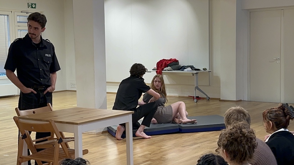 An actor wearing a police uniform stands with his hands in his pockets while two other actors crouch next to each other on mats on the floor in mid-performance of 'Faith Hope Charity'. Members of the audience are seen on the right side of the photo, watching the performance. 